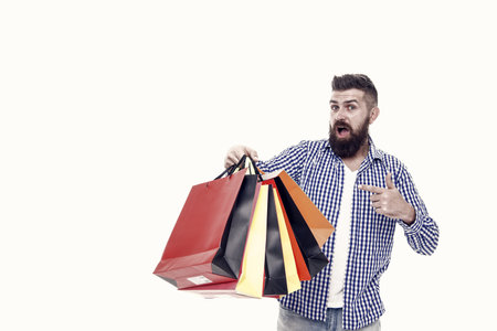 Bearded man pointing finger at shopping bags after seasonal sale isolated on white. Surprised hipster buying at sale price. Black Friday and cyber monday sale, copy spaceの写真素材