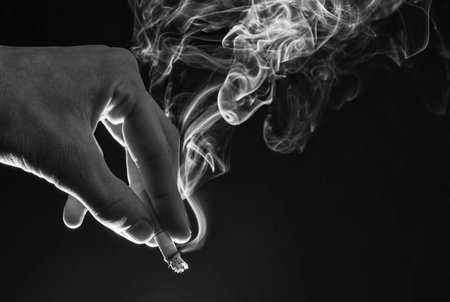 Male hand hold cigarette butt with smoke swirls dark background, nicotine addictionの写真素材