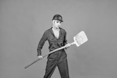 woman hard worker in protective helmet and boilersuit hold shovel on orange background, labour dayの写真素材
