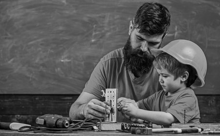 Mens work concept. Boy, child in protective helmet makes by hand, repairing, does crafts with dad. Father with beard and little son in classroom teaching to use tools, chalkboard on backgroundの写真素材