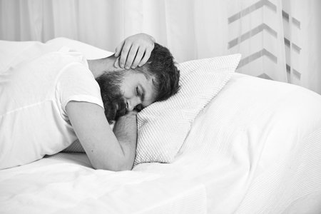 Man in shirt laying on bed covering ears with hands, white curtain on background. Wake up call concept. Macho with beard and mustache refuses to get up. Guy on sleepy tired face keep sleepingの写真素材