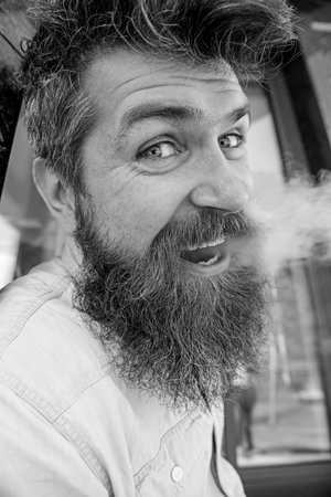 Hipster with tousled hair and gray on relaxed cheerful face with white smoke flying out of mouth. Man with beard and mustache smoking, black marble background. Smoking and habits conceptの写真素材