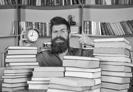 Man, scientist peeking out of piles of books with alarm clock. Teacher or student with beard studying in library. Man on happy face pointing at clock, bookshelves on background. Time flow conceptの写真素材