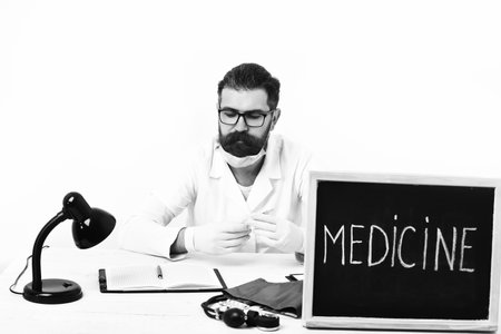Bearded man, long beard. Brutal caucasian doctor or unshaven hipster in mask, medical gown sitting at table with board Medicine inscription isolated on white studio background. Medicine concepの写真素材
