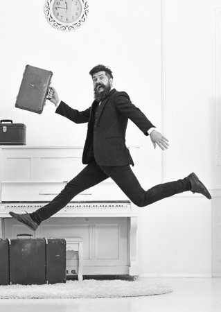 Long awaited vacation concept. Man with beard and mustache in suit carries luggage, luxury white interior background. Macho attractive, elegant on cheerful face carries vintage suitcases, jumpingの写真素材