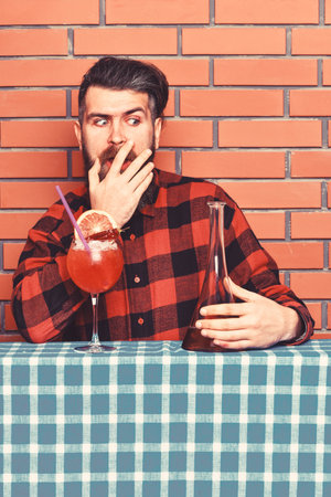 Man in checkered shirt near bottle, brick wall and blue tablecloth background. Barman with beard on shocked face holds bottle. Bartender surprised with taste of beverage. Shocking taste conceptの写真素材