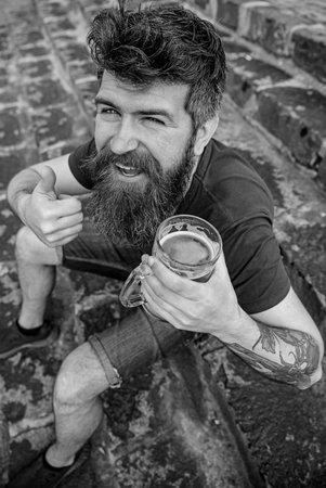 Man with beard and mustache holds glass with beer and shows thumb up, stone stairs background. Hipster on cheerful face drinking beer outdoor. Friday relax concept. Guy having rest with cold beerの写真素材