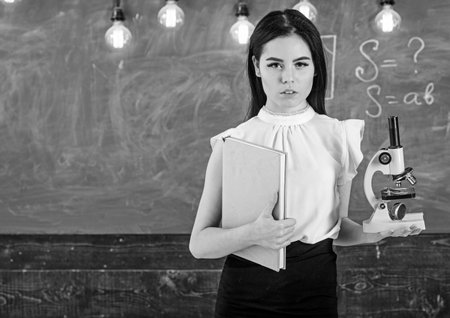 Biology concept. Teacher of biology holds book and microscope. Lady in formal wear on calm face in...