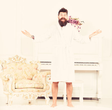 Happy morning concept. Man with beard enjoys morning while stand near piano and old fashioned armchair. Man cheerful, on surprised face in bathrobe stands in luxury hotel in morning, white backgroundの写真素材