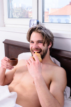 French breakfast stereotype. Guy having coffee and holds croissant while lay bed in bedroom or hotel room. Man bearded handsome guy eating breakfast in bed. Man eats croissant and drinking coffeeの写真素材