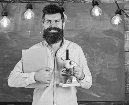 Teacher in eyeglasses holds book and microscope. Science concept. Man with beard and mustache on...