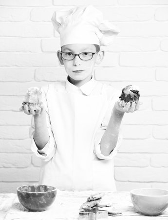 young boy cute cook chef in white uniform and hat on stained face flour with glasses standing near table with colorful bowls and holding chocolate cakes on brick wall backgroundの写真素材