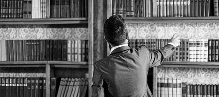 Man takes book from bookcase. Student stands in library and choose book. Guy in smart suit reads near bookcase. Study, learn, education, research, history, literature, wisdom concept. Big libraryの写真素材