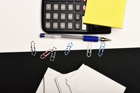 Business and work concept. Office tools on black and white background. Stationery and calculator, top view. Yellow note paper, blank business cards, pen and paper clips organised on opposite sidesの写真素材
