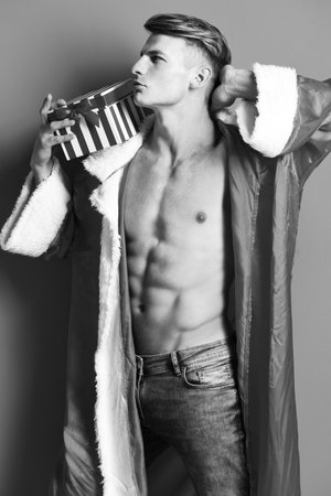 young handsome macho santa man with sexy muscular athletic strong body has bare torso and strong belly in new year sweater holding christmas or xmas present on red studio backgroundの写真素材