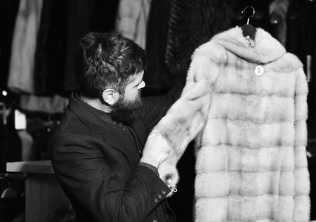 Guy holds beige furry coat in shop with fur on background. Macho with stylish appearance with mink fur in store.の写真素材