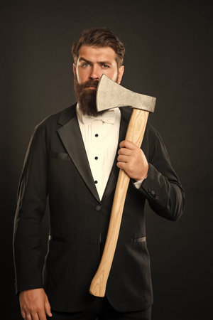 Serious bearded man in formal suit holding hatchet at unshaven face dark background, barbershopの写真素材