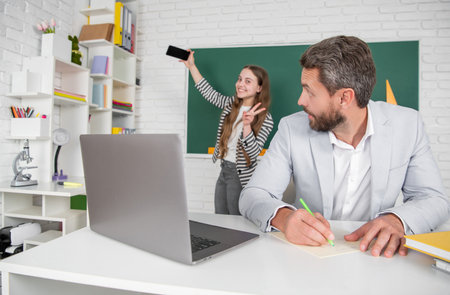 amazed school teacher in classroom with selective focus of kid making selfie at blackboard