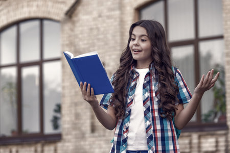 Happy kid with long wavy hair in casual style read library book preparing for school lesson outdoors, literatureの写真素材