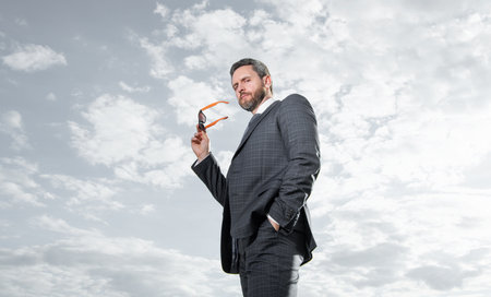 Businessman bottom view. Businessman in suit holding sunglasses. Grizzled businessman sky background