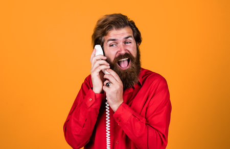 communication. bearded man wear red shirt. casual male answering call. concept of conversation. man secretary or assistant speak on retro phone. brutal man with telephoneの写真素材