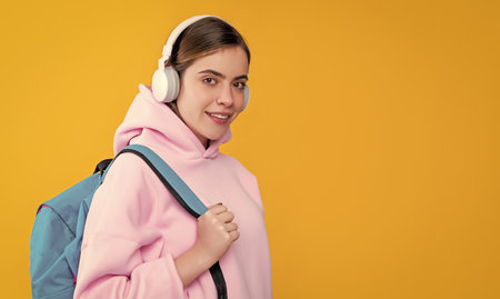 positive student girl in headphones with backpack on yellow background