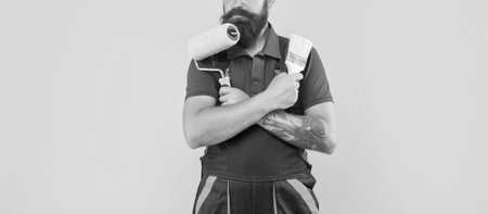 cropped man in work clothes hold paint roller and brush on yellow backgroundの写真素材