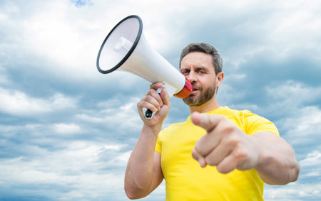 man in yellow shirt shout in loudspeaker on sky background. advertiseの写真素材