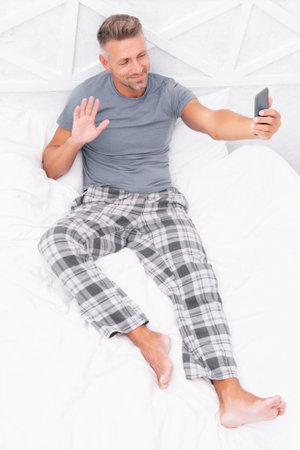 video call man with phone in bed gesturing hello. photo of video call man with phone at home.の写真素材