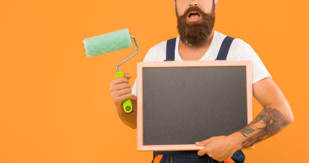 cropped man renovator hold paint roller and blackboard with copy space on yellow background.の写真素材