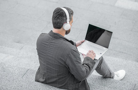 back view of man webinar online outside. photo of man webinar online on laptop with headphones. man webinar online outdoor. man webinar online in the street.の写真素材