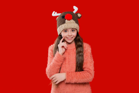 Winter child wardrobe. Teenage girl in Xmas hat isolated on red. Winter fashion for teenage style. Girl in knitted hat in winter season. Childhood lifestyle. Colorful earflap beanieの写真素材