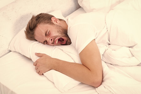 Yawning early in the morning. Asleep young man sleeping. Resting peacefully in comfortable bed. Lying with closed eyes. Man sleeping at night. Sleepy man lying on bed sleeping at white bedroomの写真素材
