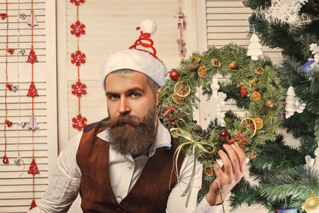 Man with moustache hold Xmas wreath as New Year decoration sitting in Santa Claus Christmas hat at homeの写真素材