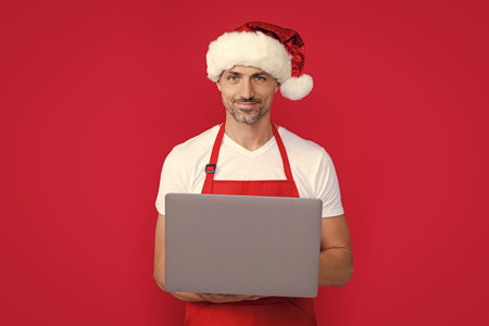 mature man in new year santa hat and red apron hold laptopの写真素材