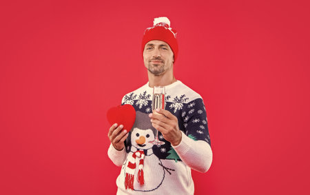 smiling man in christmas hat with champagne and heart. christmas man isolated on red background.の写真素材