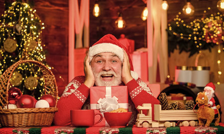 Joyful atmosphere. Senior joyful man Santa claus drinking tea. Winter holiday. Joyful grandpa with gift. Dreamy grandfather in Santa hat. New year. Cozy home. Hot beverage and cookies. Christmas eveの写真素材