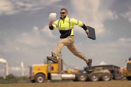 Supervisor jumping. Excited builder construction worker jumping at trucks. Man engineer running to success. Builder man run fast to work. Engineer supervisor running outdoor. Project successの写真素材