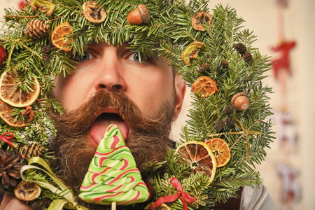 New Year holiday. Man with moustache hold Xmas wreath. Happy New Year. Christmas wreath decoration. Xmas decoration for home. Christmas man eat candy. Bearded man at Christmas. Copy spaceの写真素材