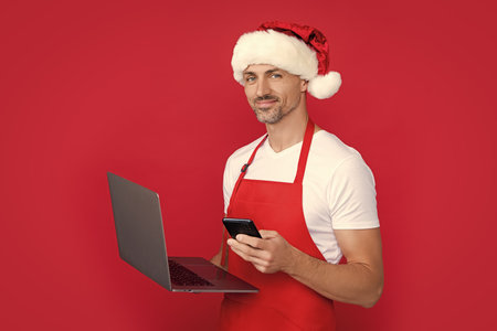 mature man in christmas santa hat and red apron use laptop with smartphoneの写真素材