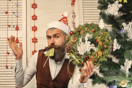 Bearded man at Christmas. New Year holiday. Man with beard hold Xmas wreath. Happy New Year. Christmas wreath decoration. Xmas decoration for home. Christmas man eat candyの写真素材