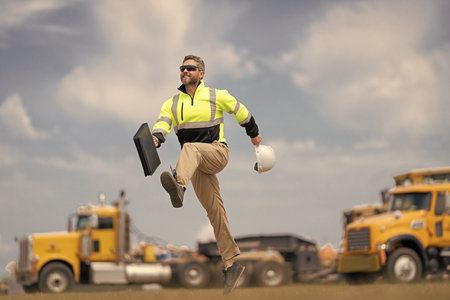 Builder man run fast to work. Engineer supervisor running outdoor. Supervisor jumping. Excited builder construction worker jumping at trucks. Man engineer running to success. End of working dayの写真素材