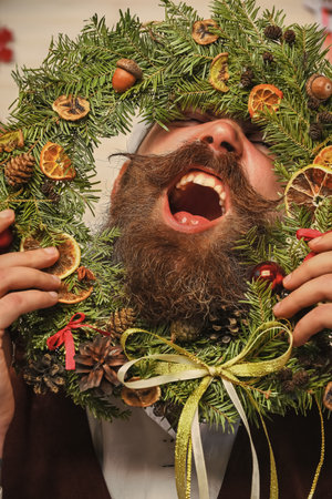 Decorated Christmas man. Bearded man at Christmas. New Year holiday. Man with moustache hold Xmas wreath. Happy New 2025 Year. Christmas wreath decoration. Xmas decoration for homeの写真素材