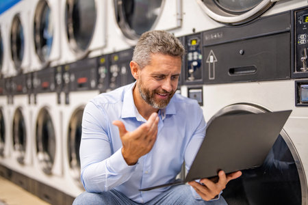 Laundry overload for busy businessman. Busy man working laptop at laundry. Overloaded employee multitasking laundry and job. Tired worker dealing laundry and deadlines. Effective planning in scheduleの写真素材