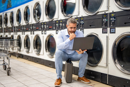Tired worker dealing laundry and deadlines. Laundry overload for busy businessman. Busy man working on laptop at laundry. Overloaded employee multitasking laundry and job. Work-life balanceの写真素材