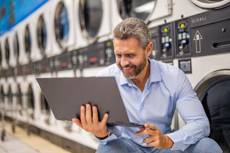 Busy man working on laptop at laundry. Overloaded employee multitasking laundry and job. Tired worker dealing laundry and deadlines. Laundry overload for busy businessman. Overloaded scheduleの写真素材