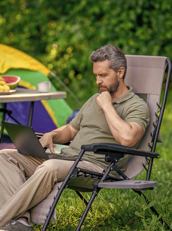 Businessman working outdoor. Man having camp picnic in forest with tent. Summer camping retreat. Business man typing on laptop at camping. Business man working online. Businessman freelancing onlineの写真素材