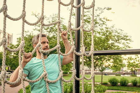 Man climbing net at obstacle course. Muscular man training at outdoor gym. Sport workout. Man sportsman do climbing exercise. Muscular strong athlete climbing net. Net climb. Copy spaceの写真素材
