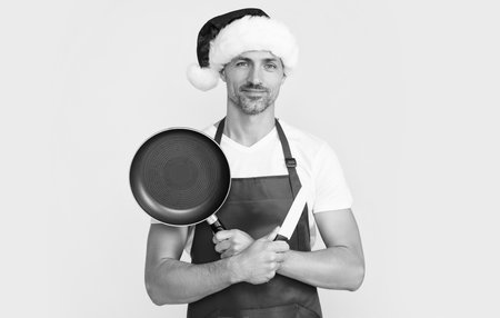 cheerful mature christmas man in red santa hat and apron hold frying pan and kitchen knifeの写真素材