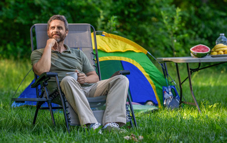 Man drink morning camp coffee in forest. Man hiker relax outdoor. Summer camp and nature vacation. Man enjoying camping. Camp adventure. Hiking concept. Camping tent. Coffee cup. Copy spaceの写真素材